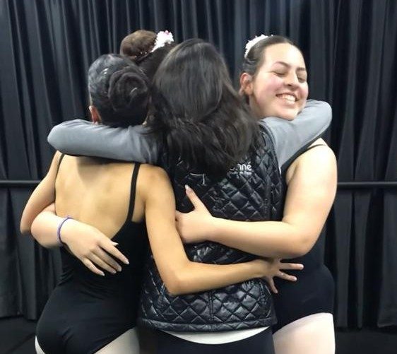 Quatro meninas, todas vestindo roupas de balé, estão abraçadas em um estúdio de dança. Elas estão sorrindo e se mostram próximas, com suas costas voltadas para a câmera. As três meninas à esquerda usam trajes de balé preto e meias brancas, enquanto a quarta, no meio, veste uma jaqueta preta sobre o traje de balé. O fundo é de cortinas escuras, típicas de um ambiente de estúdio.