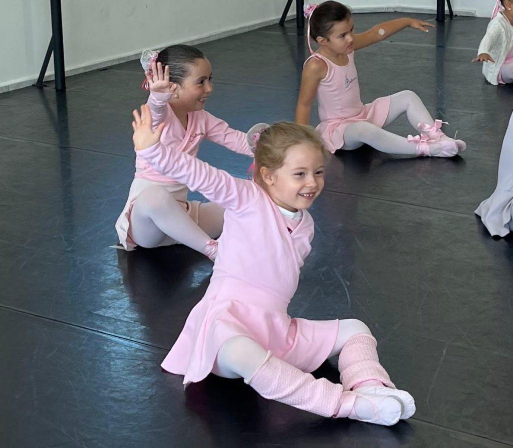 Três meninas pequenas vestidas com roupas de balé rosa praticam uma coreografia sentadas no chão de um estúdio de dança. Elas estão sorrindo e com um dos braços erguidos, demonstrando entusiasmo e alegria. Ao fundo, outras crianças também participam da aula. O ambiente tem piso preto próprio para dança e paredes brancas.
