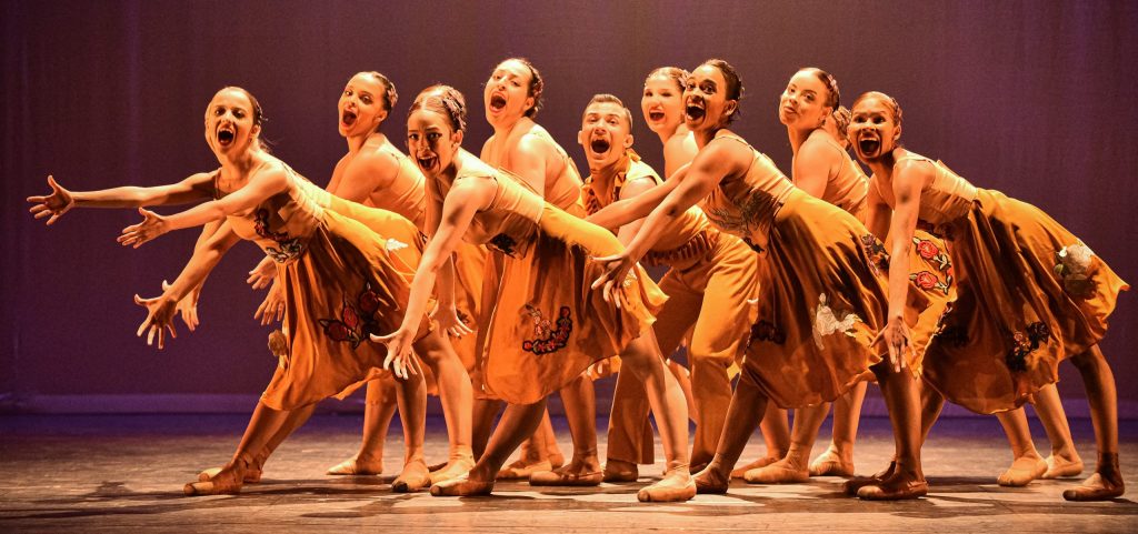 Grupo de dançarinas em um palco, vestindo vestidos amarelos com detalhes coloridos. Elas estão em uma performance enérgica, com expressões faciais intensas e braços estendidos para frente. O fundo é iluminado com luzes suaves, criando um ambiente dramático para a apresentação.
