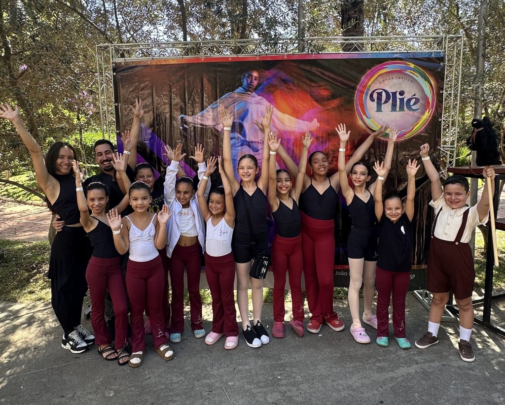 Grupo de crianças e professores posando com os braços levantados em frente a um banner colorido com o logo "Plié Dance Festival". As crianças estão usando roupas de dança vermelhas e pretas, com algumas usando tênis e chinelos. Todos sorriem e se mostram animados.