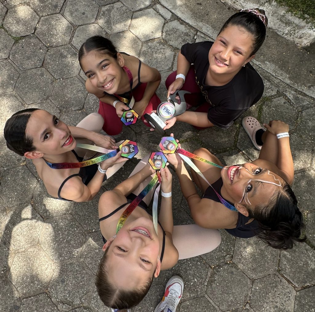 Um grupo de cinco dançarinas sorrindo enquanto seguram suas medalhas de premiação. Elas estão reunidas ao ar livre, com roupas de dança e medalhas coloridas ao redor de seus pescoços, celebrando sua conquista.