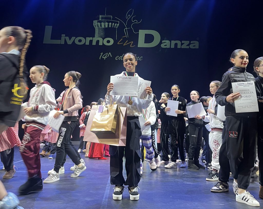 Uma dançarina sorrindo e segurando um diploma e uma medalha de ouro no palco de uma competição de dança. Ao fundo, outros participantes estão recebendo seus prêmios. O evento é parte do "Livorno in Danza", um festival de dança.
