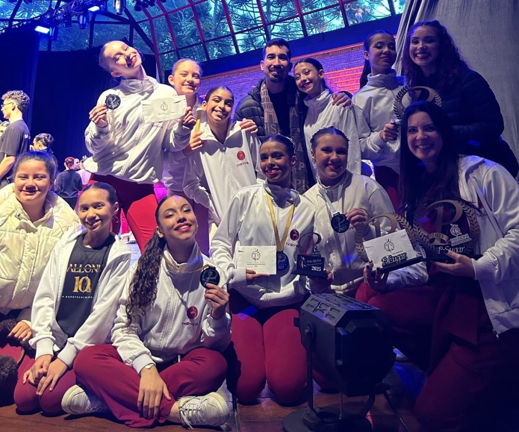 Um grupo de dançarinas e instrutores posando felizes no palco, todos usando roupas de aquecimento brancas e calças vermelhas. Elas seguram vários troféus e medalhas, comemorando suas vitórias em uma competição de dança.