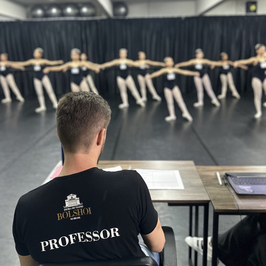 Vista de um professor de balé, com a camiseta escrita 'Escola do Teatro Bolshoi no Brasil' e 'PROFESSOR', observando um grupo de bailarinas durante o exame. As bailarinas estão alinhadas à frente, com os braços estendidos para os lados, e usam trajes pretos e sapatilhas de balé. O professor está sentado à mesa, com papéis e um computador à sua frente.