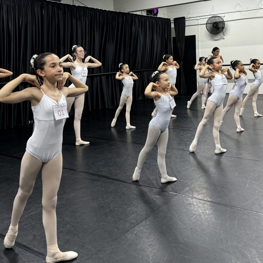 Grupo de crianças fazendo o exame de balé, todas vestindo trajes brancos e sapatilhas. Elas estão alinhadas e com as mãos nos ombros, praticando a posição sob a supervisão de uma instrutora. Cada criança tem um número de identificação visível em seus trajes.